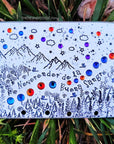 Silver toned rectangle is lying in grass. It's decorated with a mountain scene, set with purple and orange toned crystals, and reads "Neverender de la Beuna Sangre". 