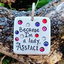 Load image into Gallery viewer, A silvered colored pet tag that says &quot;Because I&#39;m a lady, assface&quot; in black letters with red, blue, white, and pink crystals is on a wood background. 
