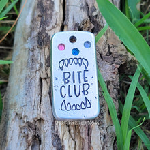 Load image into Gallery viewer, Silver colored rectangle pet tag with &quot;Bite Club&quot;, fangs, and pink, purple, and blue crystals on a wood background. 
