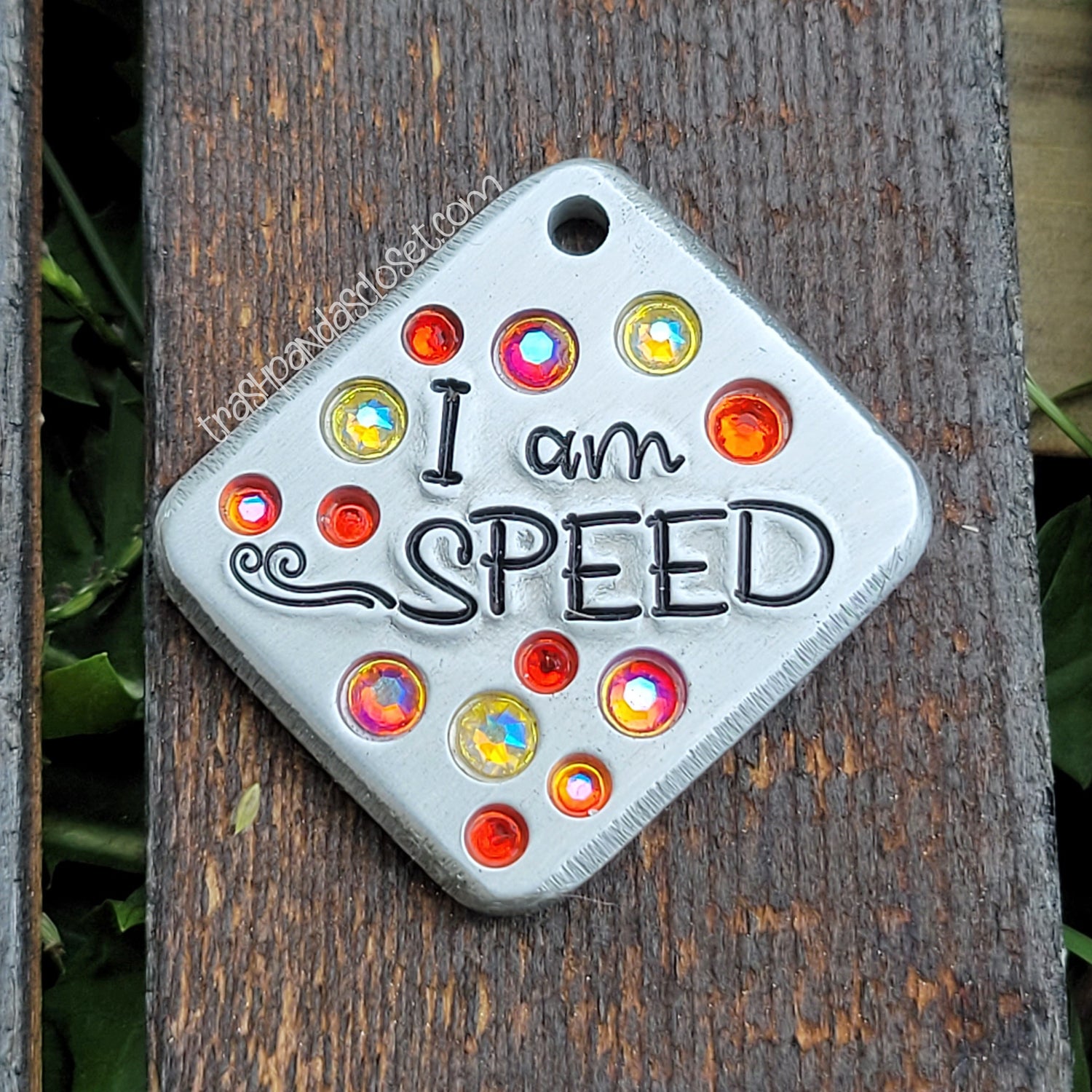 Silver colored pet tag with the words "I am speed" a whoosh design, and orange and yellow crystals lying on a wood slat background. 