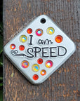 Silver colored pet tag with the words "I am speed" a whoosh design, and orange and yellow crystals lying on a wood slat background. 