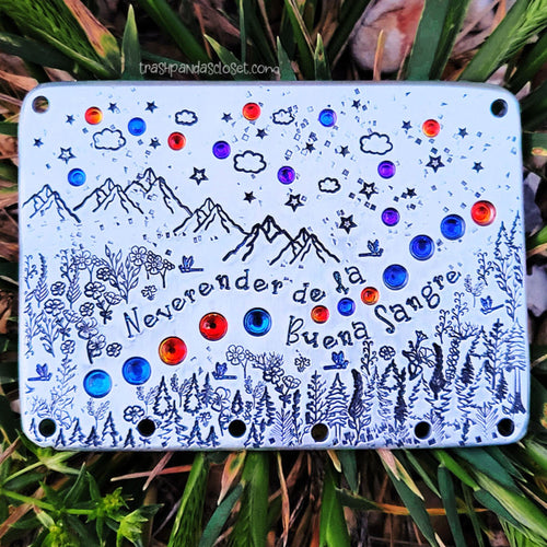 Silver toned rectangle is lying in grass. It's decorated with a mountain scene, set with purple and orange toned crystals, and reads 